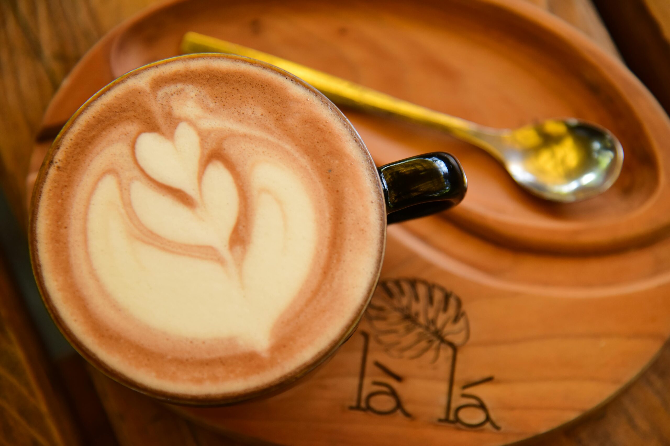 Intricate latte art coffee on a wooden tray with a spoon, in a cozy cafe setting.